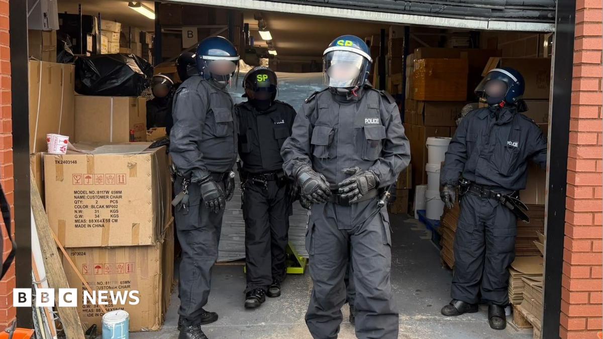 A group of four police officers in all-black uniforms and with helmets and masks are standing in a warehouse section of a red brick building.