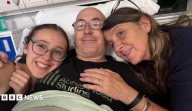 Three people are in a selfie in a hospital bed. On the left, there is a girl with her dark hair behind her back. She is wearing dark green rimmed glasses and a black T-shirt. In the middle, there is a man who is lying in the bed. He is wearing glasses and a black T-shirt that says 'Abbey Road Studios'. On the right, there is a woman with brown shoulder length hair and a dark blue jumper.