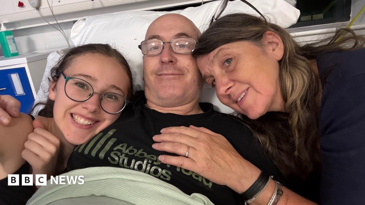 Three people are in a selfie in a hospital bed. On the left, there is a girl with her dark hair behind her back. She is wearing dark green rimmed glasses and a black T-shirt. In the middle, there is a man who is lying in the bed. He is wearing glasses and a black T-shirt that says 'Abbey Road Studios'. On the right, there is a woman with brown shoulder length hair and a dark blue jumper.