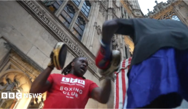 A person wearing a red London Fight Factory boxing club T-shirt holds focus pads while another person throws punches during an outdoor boxing session at Parliament