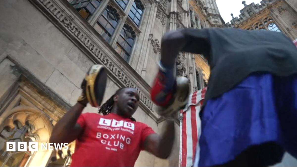 A person wearing a red London Fight Factory boxing club T-shirt holds focus pads while another person throws punches during an outdoor boxing session at Parliament
