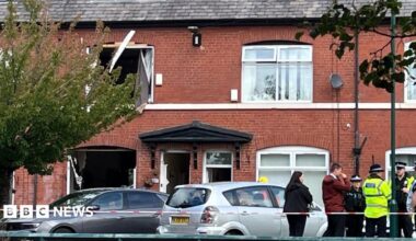Image shows police and civilians standing behind a line of police tape, with a house which has had its windows blown out in the background.