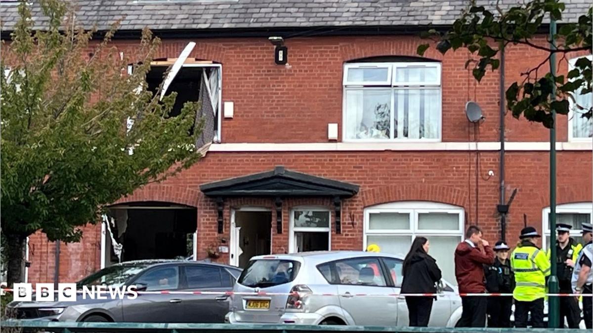Image shows police and civilians standing behind a line of police tape, with a house which has had its windows blown out in the background.