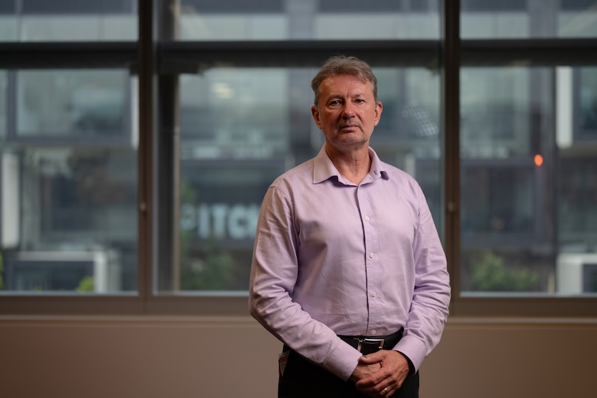 A man (Ty Christopher) wearing a lilac collared shirt looking serious in an office.