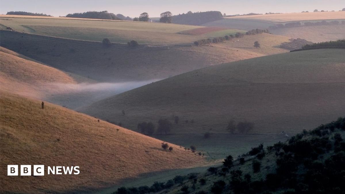 A series of slopes overlooking a valley with mist at the bottom. Trees are dotted around the landscape.