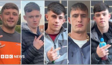 Five young men in their late teens and early 20s. They all have similar haircuts, with a fringe and faded back and sides. Four of them are gesturing with their fingers. Three of them extend their thumb, index, and pinky fingers - leaving their middle and ring finger tucked down. One points with his index and middle finger whilst holding a Lucozade bottle. They are all dressed in casual clothing.