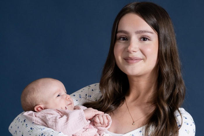 DSC03485 Sarah smiles at the camera holding baby Leyla, stood against a neutral deep blue background in key art for Emmerdale
