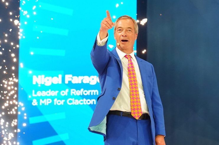 Nigel Farage speaking at the Reform UK conference, pointing upwards with a confident expression.