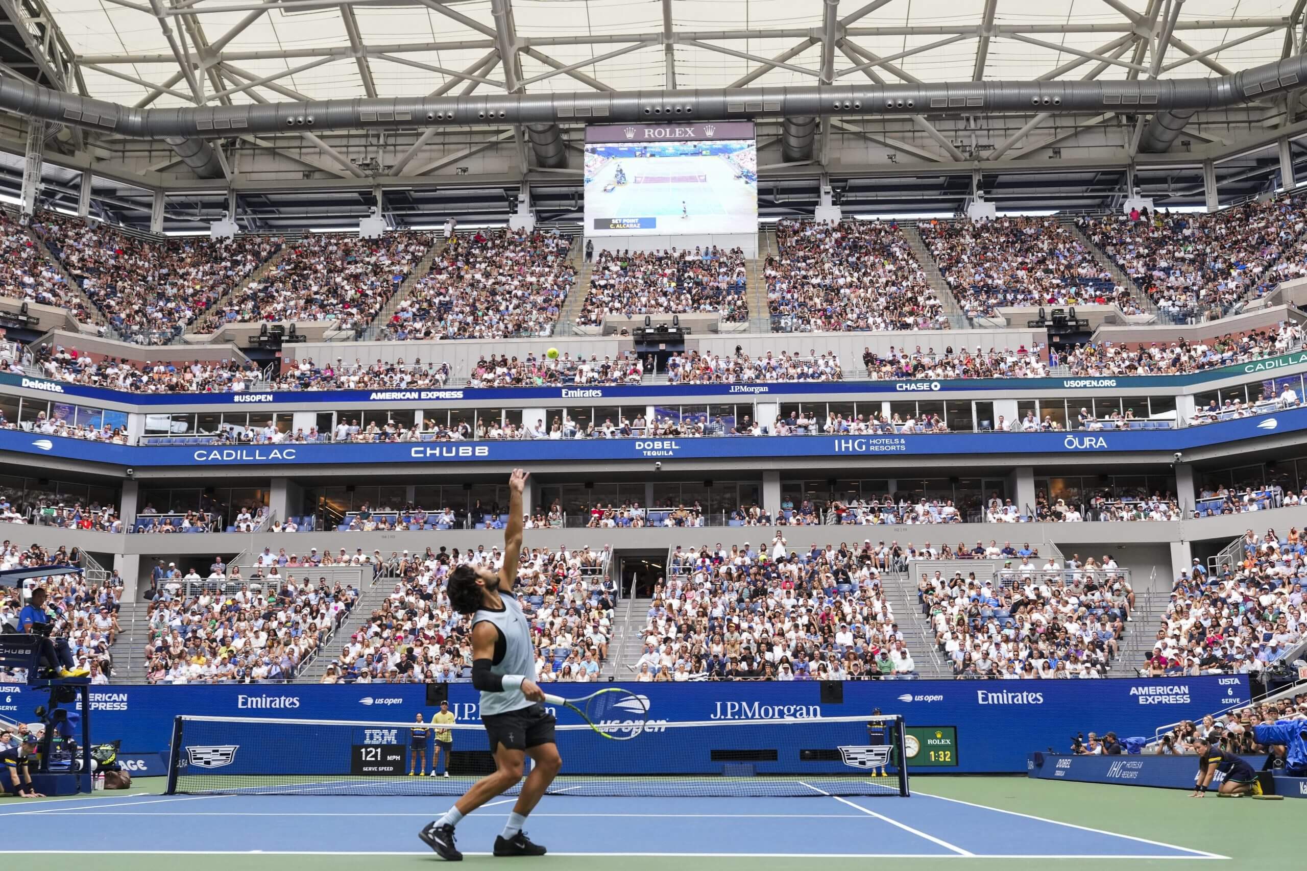 A wide view of Carlos Alcaraz tossing the ball up to serve, with a view of fans in the stadium in the background.