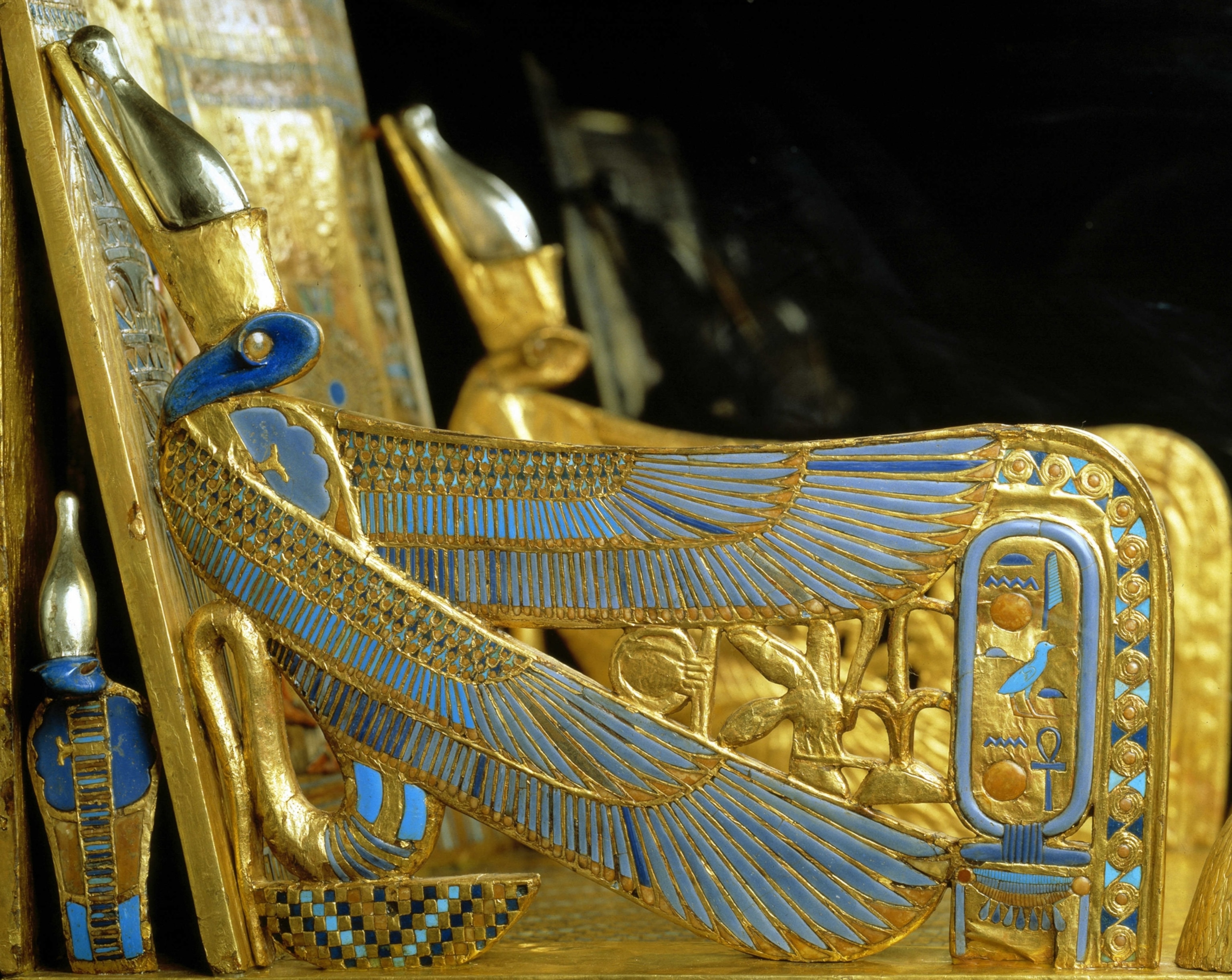 A close-up of an ancient Egyptian gold-and-blue decorative artifact featuring a stylized bird with outstretched wings and hieroglyphic symbols.