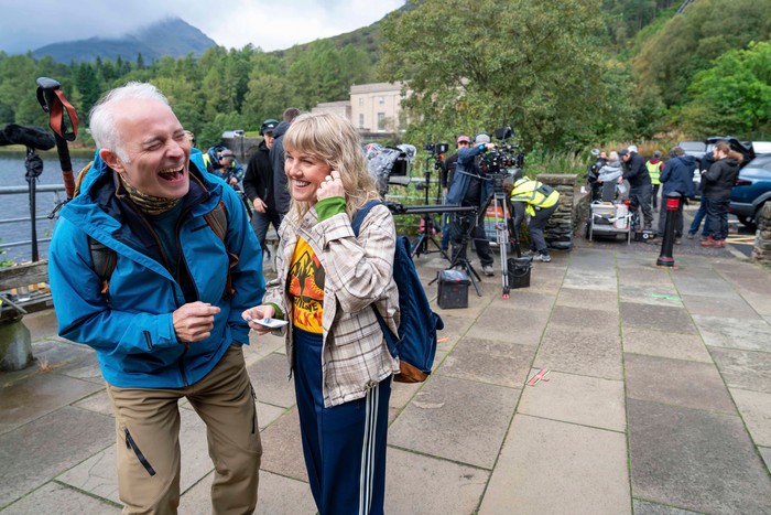 Mark Bonnar and Ashley Jensen candidly laughing on set of Rambling wearing hiking gear with production crew visible behind them.