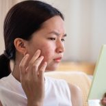 Portrait of Asian woman worry about her face when she saw the problem of acne inflammation and scar by the mini mirror.