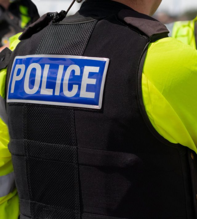 Close-up of 'POLICE' marking written on the back of a hi-visibility stab proof vest worn by a trio of police officers at the scene of an incident. Officers enforce the law, establish public order and protect citizens.
