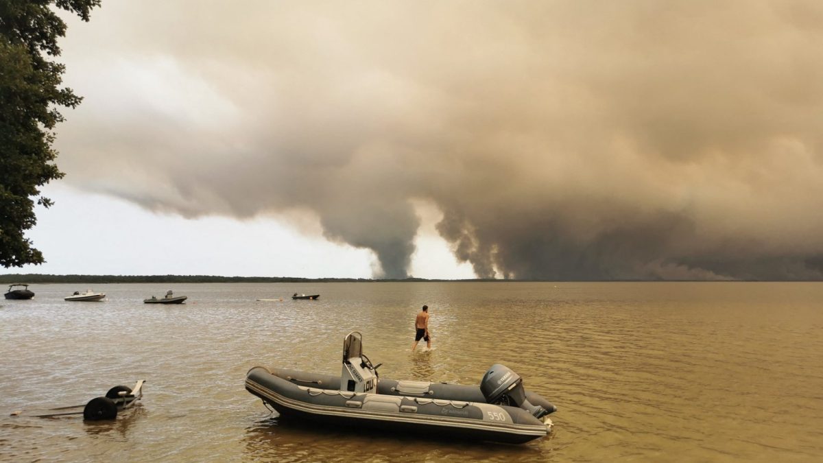 CORRECTION / A beachgoer walks in Lake Lacanau, also called Etang de Lacanau (Lacanau Pond) from the beach of Moutchic in Lacanau, southwestern France on July 24, 2026, as clouds of smoke rise into the sky from a wildfire. The fire raging in the northern part of the Arcachon Basin in Gironde, southwest France, has swept through more than 3,400 hectares, according to the latest figures released by the prefecture. More than 20,000 residents and tourists have been evacuated from this part already devastated by flames in 2022. Scientists have long warned that climate change caused by human use of fossil fuels will make periods of drought more intense and longer-lasting, creating the ideal conditions for wildfires. (Photo by Maximilien LAMY / AFP via Getty Images) / 