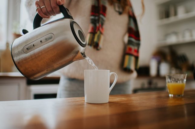 A woman pours boiling water into a white mug from a silver kettle.