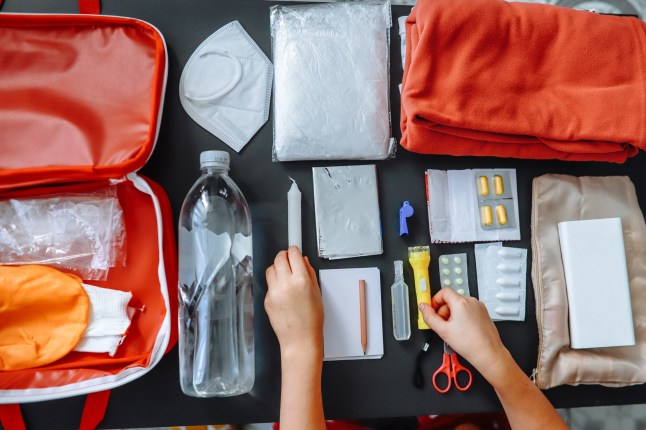 Close-up of women's hands packing a flashlight in doomsday prepping on the table at home. Includes warm clothing, flashlight, power bank, thermal blanket, medicines, water, and other emergency supplies for disasters.