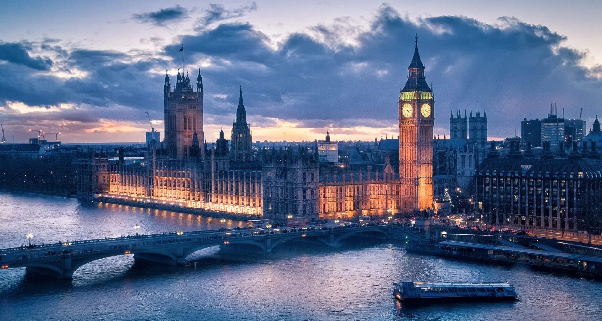 A view of the Houses of Parliament and Big Ben in London, illuminated at sunset with the River Thames in the foreground.