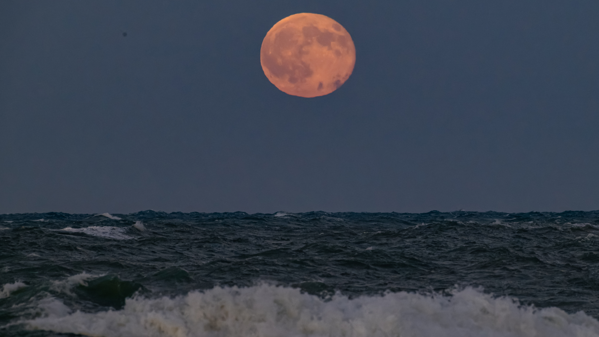 An orange full moon shines in a dark sky above crashing ocean waves.