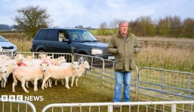 Jeremy Clarkson stood in front of sheep in a holding pen with his hands in his pockets on his farm, a land rover is parked behind him.