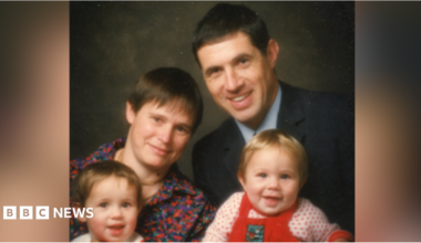 A family photograph of Helen and Clive with their two young children. Helen has short brown hair and wears a red and blue patterned top. Clive wears a suit with a pale blue shirt. Helen holds a toddler who is wearing a red jumper while Clive holds a baby wearing red dungarees.