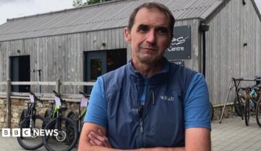A man looks at the camera with a slight smile. There are bicycles behind him and he is standing outside.