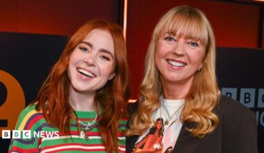 Angela Scanlon and Sara Cox standing and smiling together in front of the BBC Radio 2 logo