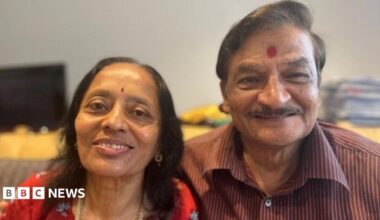 A hindu couple are looking straight at the camera and smiling. The woman is wearing a red top with flowers on it, she has thin black hair just past her shoulders and she has a small red dot called a bindi in the middle of her forehead. Her husband has his arm around here and is wearing a maroon verticle striped shirt and has a large bindi on his forehead.