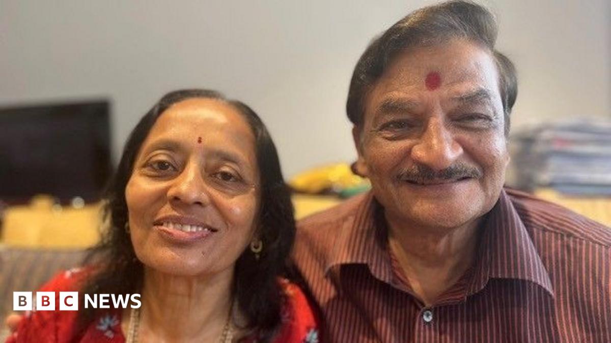 A hindu couple are looking straight at the camera and smiling. The woman is wearing a red top with flowers on it, she has thin black hair just past her shoulders and she has a small red dot called a bindi in the middle of her forehead. Her husband has his arm around here and is wearing a maroon verticle striped shirt and has a large bindi on his forehead.