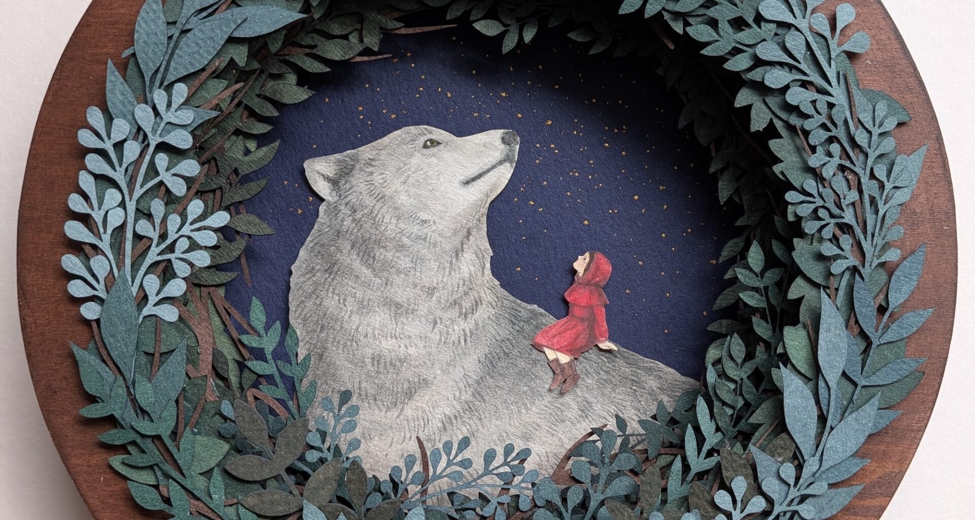 a paper collage on a round surface featuring a wreath-like arrangement of foliage around a large wolf and a young girl in a red cloak
