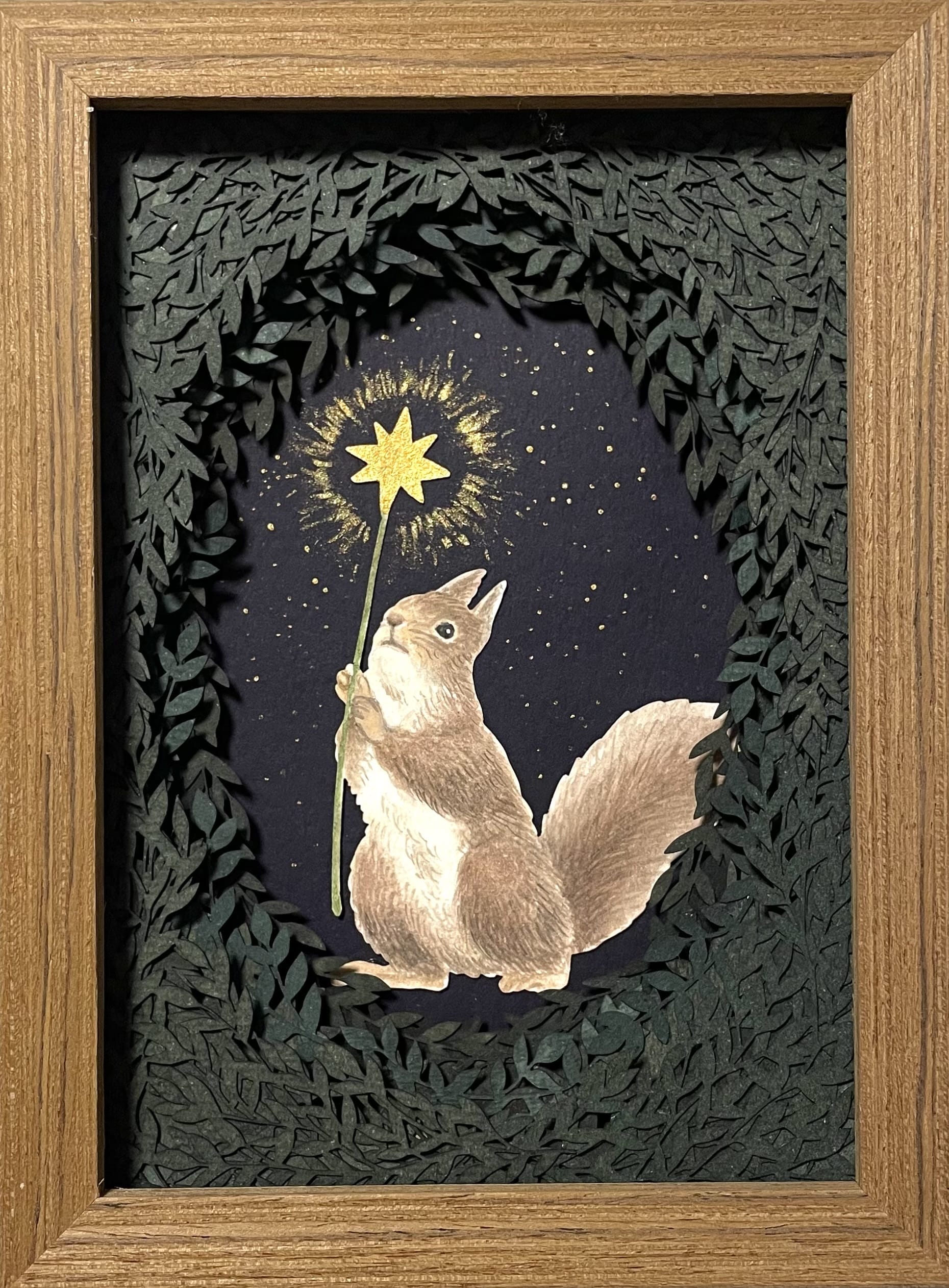 a small paper collage in a wooden frame of a squirrel with a glowing star peeking out from behind a surround of foliage