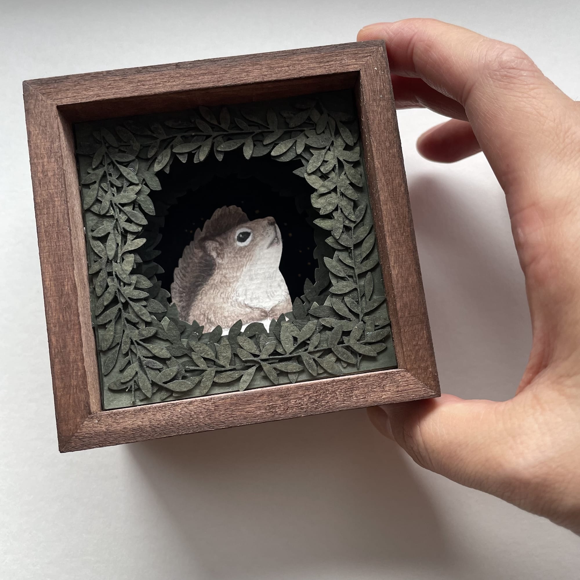 a hand holds a small paper collage in a wooden frame of a squirrel peeking out from behind a surround of foliage