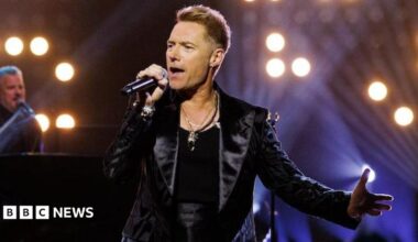 Ronan Keating performing on stage at the Riverside Studios, holding a microphone and wearing a black vest and black leather jacket