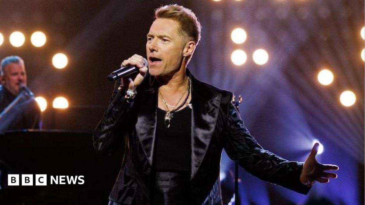 Ronan Keating performing on stage at the Riverside Studios, holding a microphone and wearing a black vest and black leather jacket