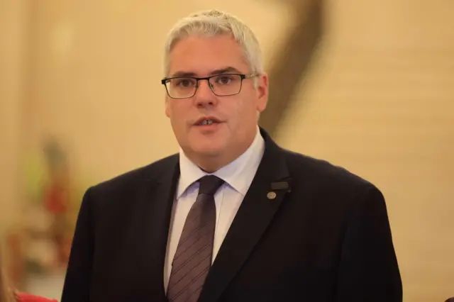 DUP leader Gavin Robinson wearing dark rimmed glasses and a black suit with white shirt and tie