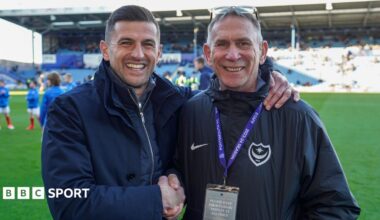 Alan Knight standing by the side of the Fratton Park pitch shaking manager John Mousinho's hand whilst Mousinho has his left arm around Knight's shoulders in embrace, both smiling