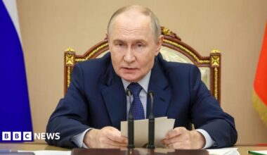Putin sits in front of a desk and microphone holding some notes
