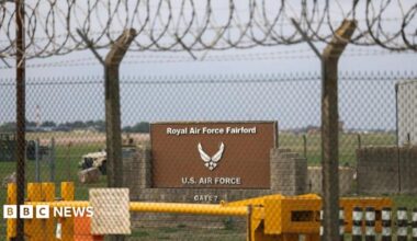 A sign stands behind a fence at the RAF Fairford airbase. Two vehicles can be seen in the background on a field behind the sign