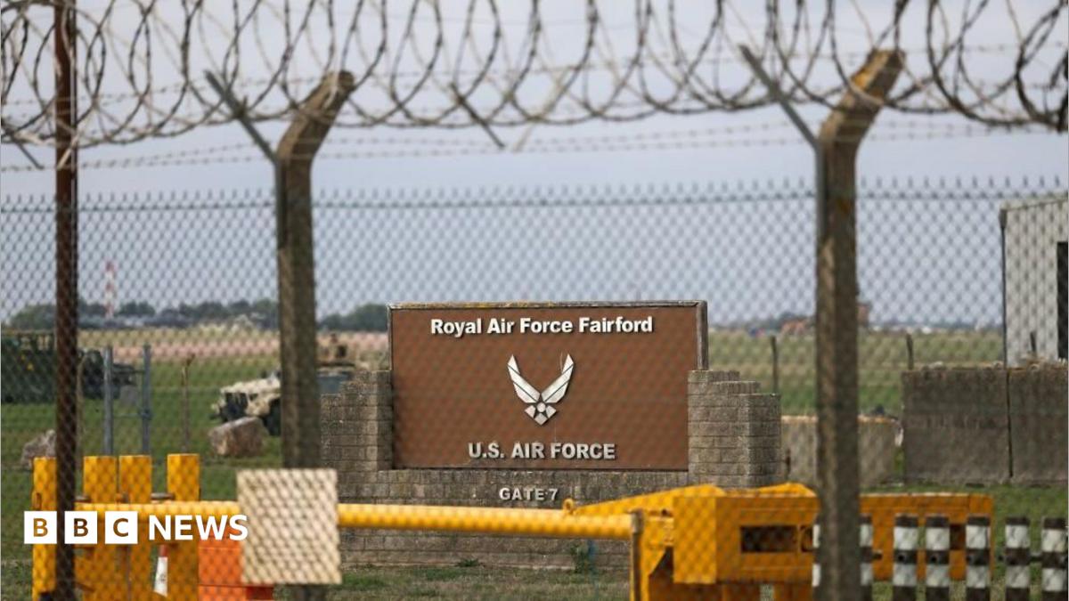 A sign stands behind a fence at the RAF Fairford airbase. Two vehicles can be seen in the background on a field behind the sign