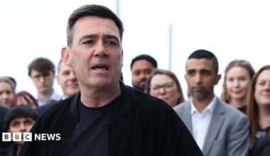 Andy Burnham is wearing a dark jacket over a dark top. He is stood in front of a crowd of people and is speaking.