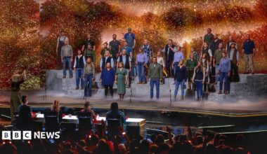 A large group of performers stands on tiered platforms on a brightly lit stage, appearing to sing together as a choir. A conductor stands at the front, guiding the performance. In front of the stage, a panel of judges sits at a table, watching. The background features warm, glowing lights and foliage-like scenery, while an audience in the foreground raises hands and watches the show.
