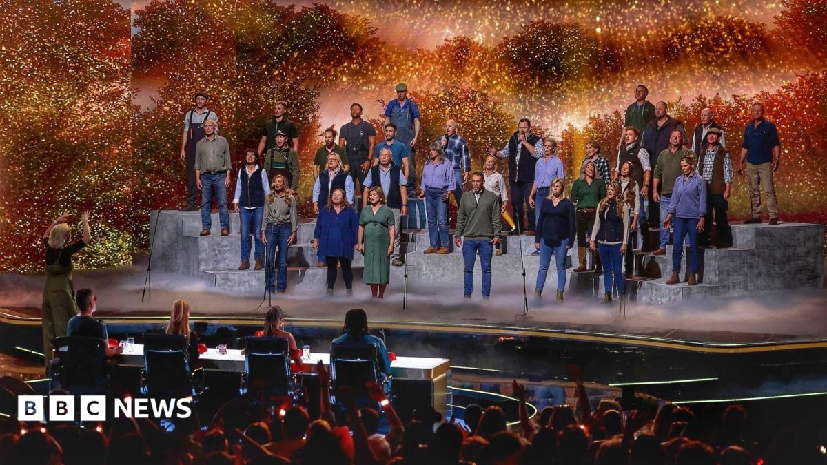 A large group of performers stands on tiered platforms on a brightly lit stage, appearing to sing together as a choir. A conductor stands at the front, guiding the performance. In front of the stage, a panel of judges sits at a table, watching. The background features warm, glowing lights and foliage-like scenery, while an audience in the foreground raises hands and watches the show.