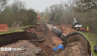 A huge sunken hole with no water in it, where a canal once was. The sides of the canal have collapsed, exposing mud and creating a large hole. Wooden fences on either side have fallen into the hole. There are two dark green canal boats sitting at the bottom of the hole, along with various debris scattered like blue tarpaulin and life rings. There are three workers in hi-vis clothing on the right hand bank, next to a white digger. In the distance, on higher ground, is a huge red and yellow pulley system ready to drag the boats out of the hole