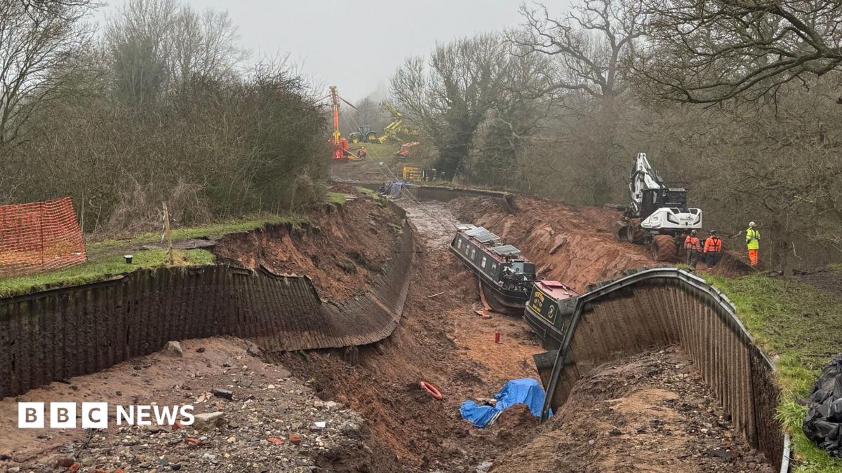 A huge sunken hole with no water in it, where a canal once was. The sides of the canal have collapsed, exposing mud and creating a large hole. Wooden fences on either side have fallen into the hole. There are two dark green canal boats sitting at the bottom of the hole, along with various debris scattered like blue tarpaulin and life rings. There are three workers in hi-vis clothing on the right hand bank, next to a white digger. In the distance, on higher ground, is a huge red and yellow pulley system ready to drag the boats out of the hole