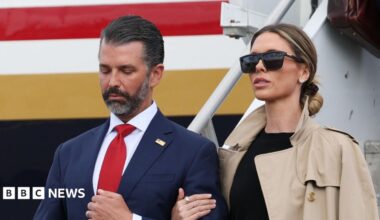Donald Trump Jr. and his wife Bettina Anderson are seen close up with a plane behind them. She is on the right in a blacktop, tan trench coat and black sunglasses. Her hair is pulled back. He is wearing a blue suit and red tie.