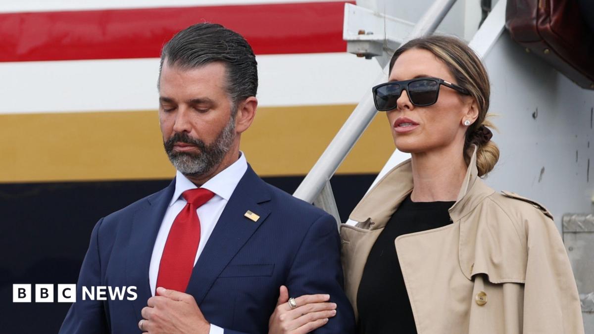 Donald Trump Jr. and his wife Bettina Anderson are seen close up with a plane behind them. She is on the right in a blacktop, tan trench coat and black sunglasses. Her hair is pulled back. He is wearing a blue suit and red tie.