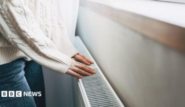 Person wearing white jumper and jeans placing their hands on a radiator.