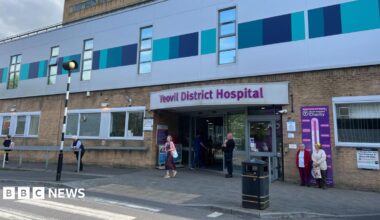 a zebra crossing leading to the main entrance of yeovil district hospital with the hospital's name in purple lettering above an automatic door with a security guard outside. The rest of the hospital building is yellow brick or clad in grey and turquoise panels