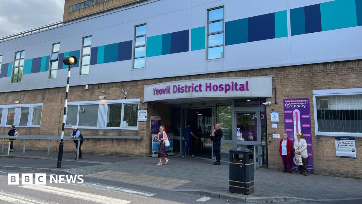 a zebra crossing leading to the main entrance of yeovil district hospital with the hospital's name in purple lettering above an automatic door with a security guard outside. The rest of the hospital building is yellow brick or clad in grey and turquoise panels