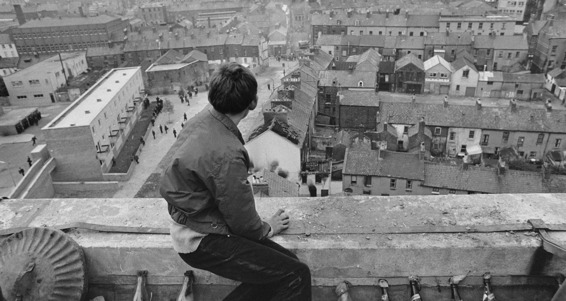 a historical black-and-white photo of a boy seated on a parapet, with rows of glass bottles filled with rags like napalm, looking down on a town