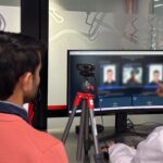 Back of a person in an orange shirt watching a large monitor showing a video conference with blurred participants; a tripod camera sits on the desk nearby.
